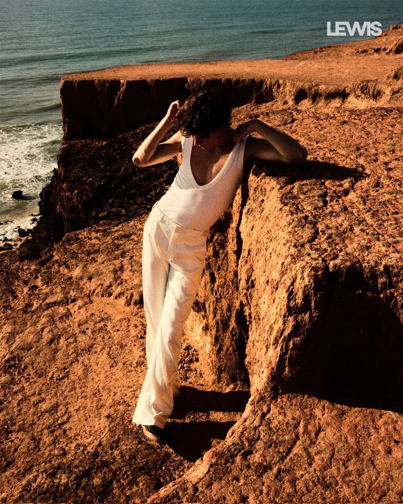 Woman in white outfit poses on a cliff overlooking the ocean. The scene features a textured cliffside and bright, natural lighting.