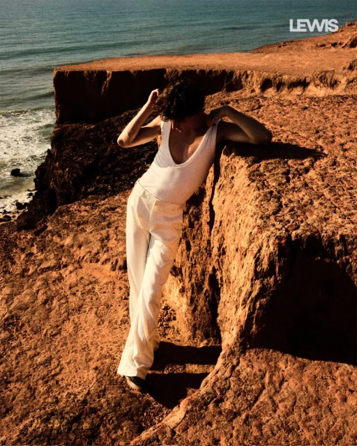Woman in white outfit poses on a cliff overlooking the ocean. The scene features a textured cliffside and bright, natural lighting.