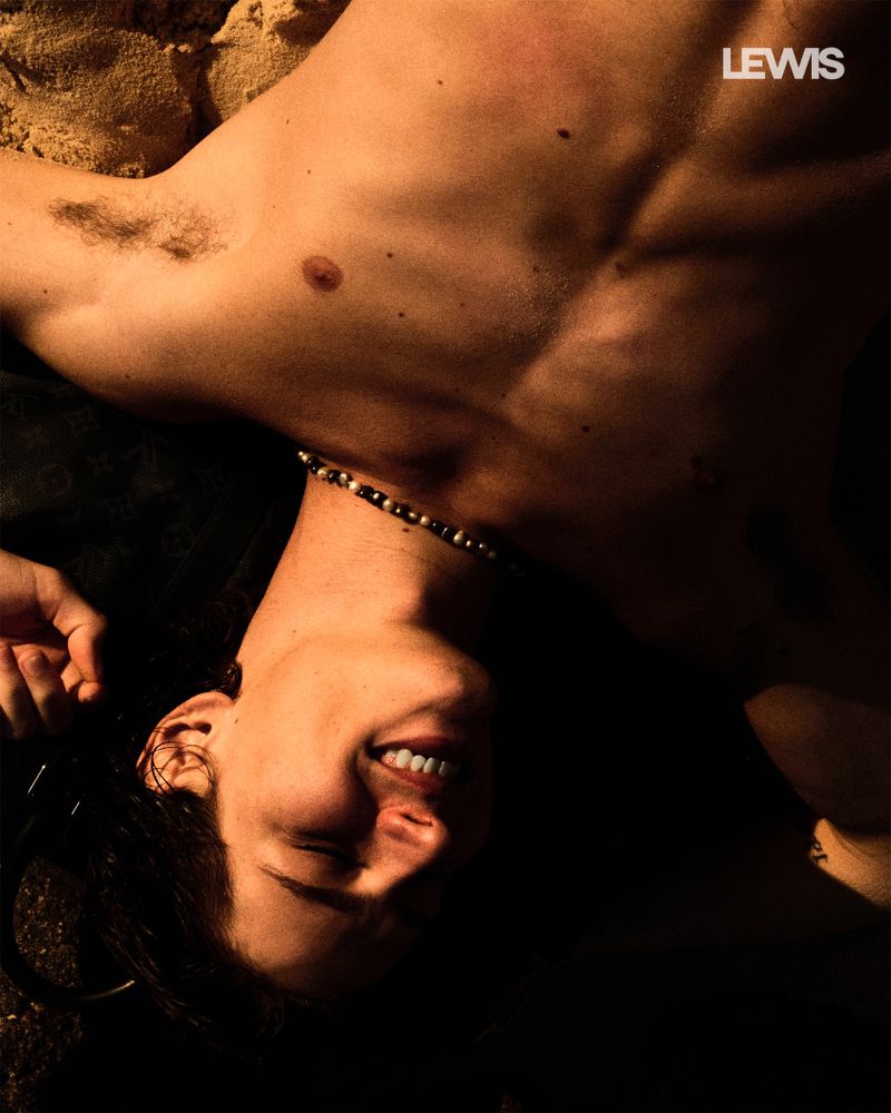 Chest and face of a shirtless adult male with a necklace laying on beach sand in soft light, smiling and looking upwards.