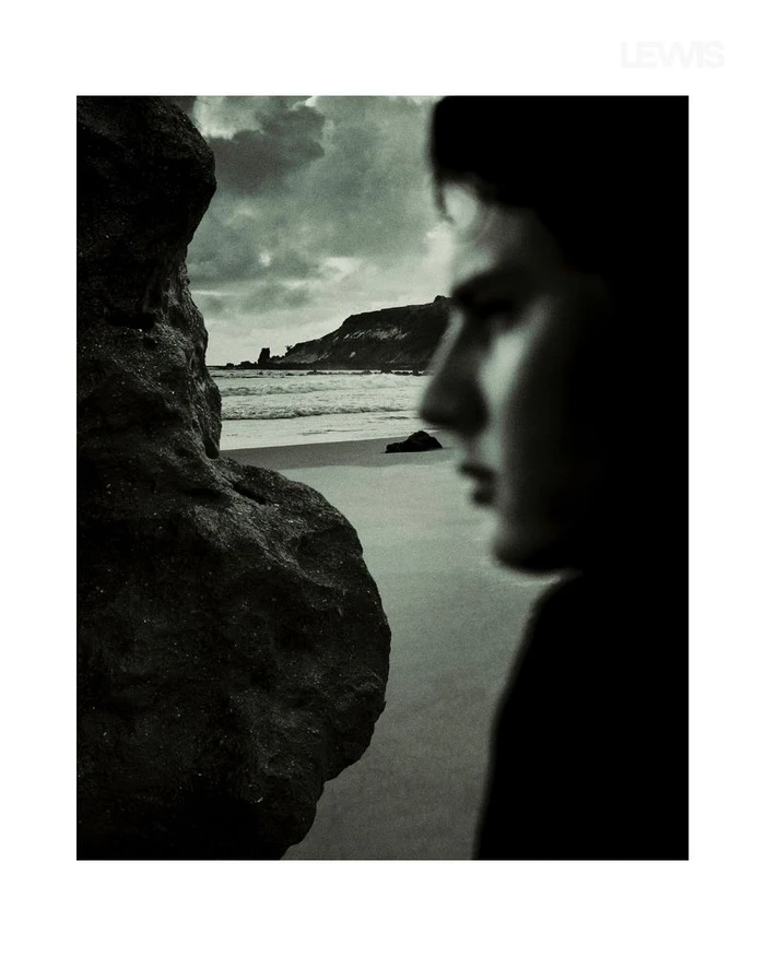 Profile of a young man looking toward a rocky coastal beach with dark, moody storm clouds in the background.