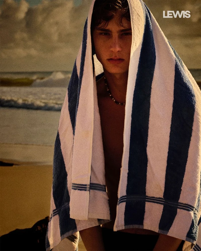 Young man with dark hair draped in a blue and white striped towel standing outdoors on a sandy beach near the ocean