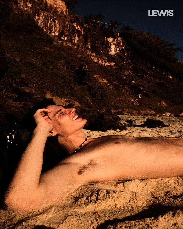 Shirtless man lying smiling on a sandy beach below a rocky cliff at sunset for Lewis Magazine