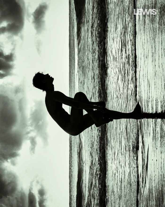 Silhouette of a man posing against a textured shoreline with a cloudy sky background in a vertical composition.