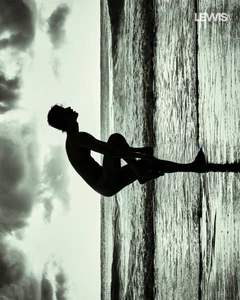Silhouette of a man posing against a textured shoreline with a cloudy sky background in a vertical composition.