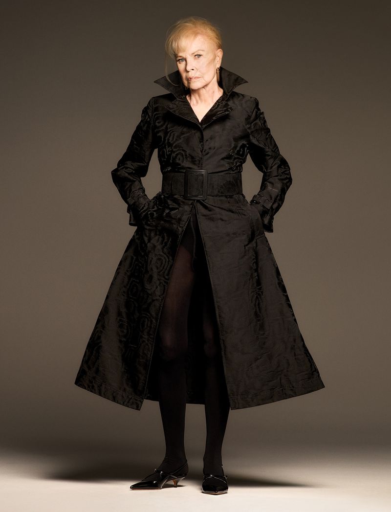 An elderly woman with blonde hair, wearing a long black patterned coat with a wide belt and black kitten heels, poses in a photo studio.