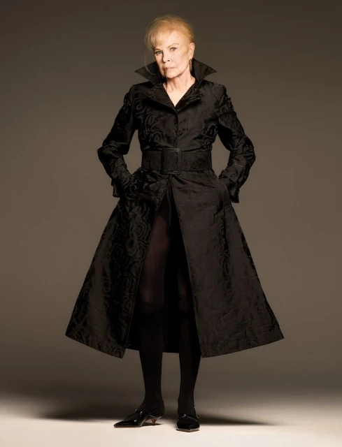 An elderly woman with blonde hair, wearing a long black patterned coat with a wide belt and black kitten heels, poses in a photo studio.