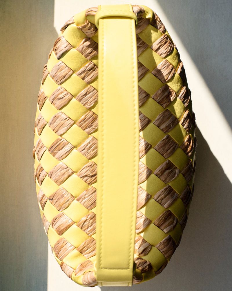 Close-up of woven handbag in yellow leather and natural raffia material with yellow leather handle, lit by bright sunlight.