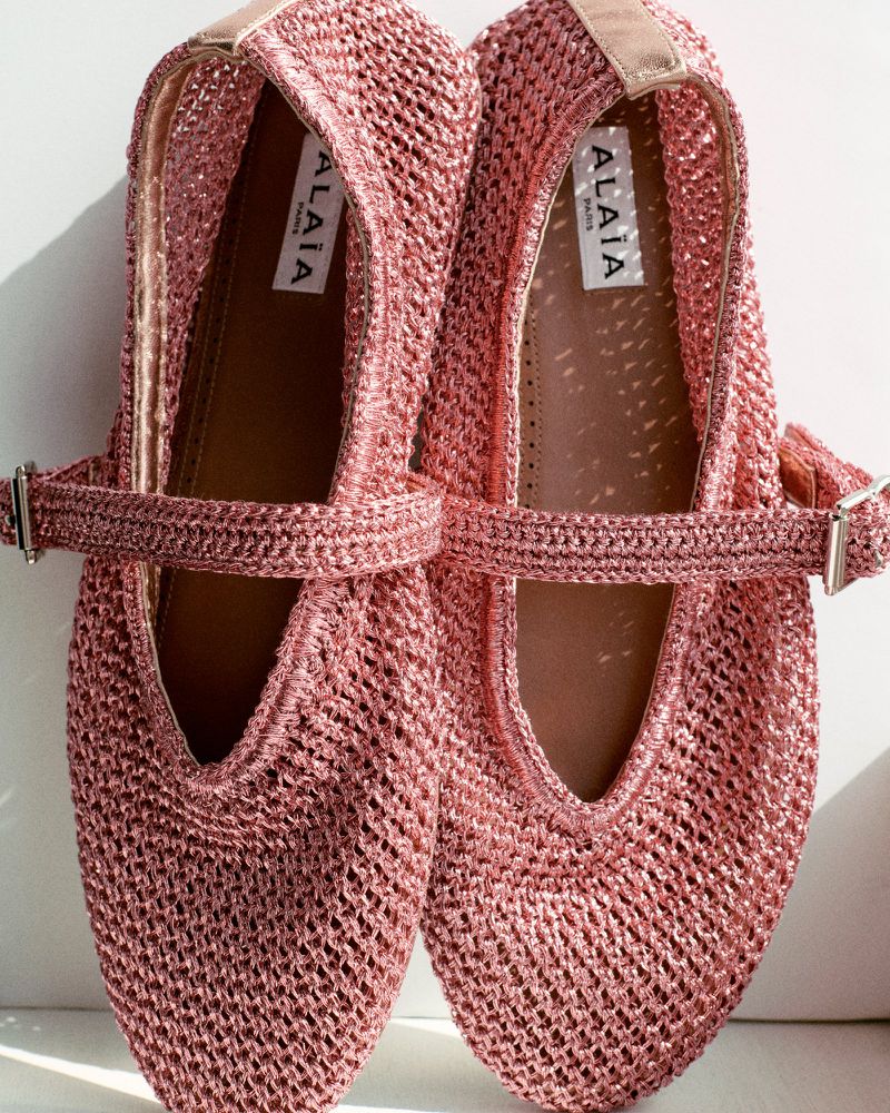 Close-up of a pair of pink, woven ballet flats with buckle straps, showcasing fashion footwear in a studio setting with bright lighting.