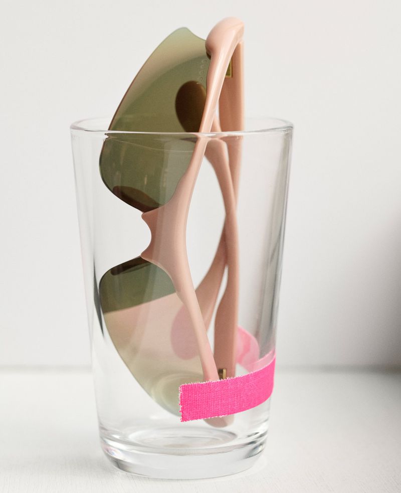 Sunglasses in a clear glass with pink tape around it against a plain white background create an interesting still life.