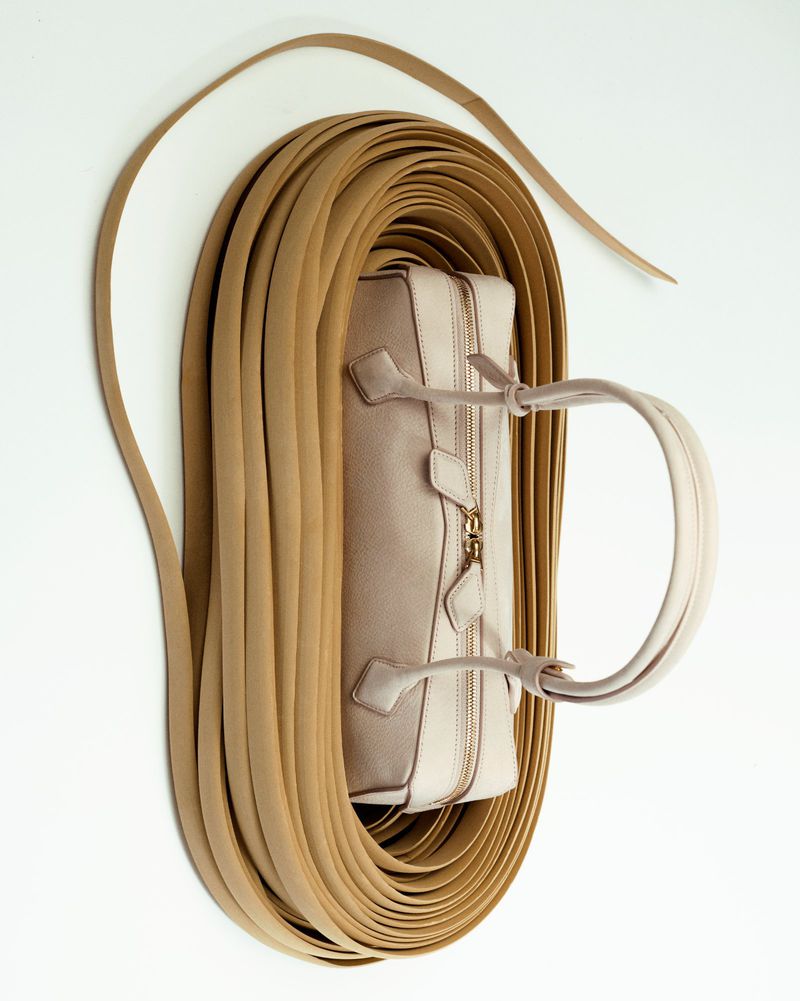 Beige leather handbag surrounded by a coil of tan leather straps on a white background, in a studio shot emphasizing materials and design.