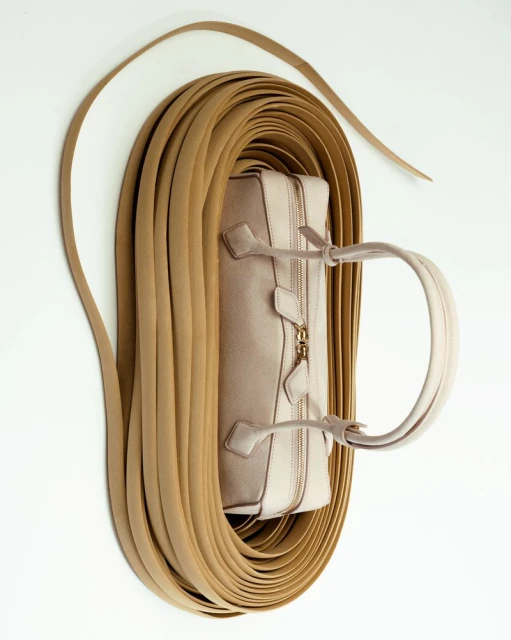 Beige leather handbag surrounded by a coil of tan leather straps on a white background, in a studio shot emphasizing materials and design.