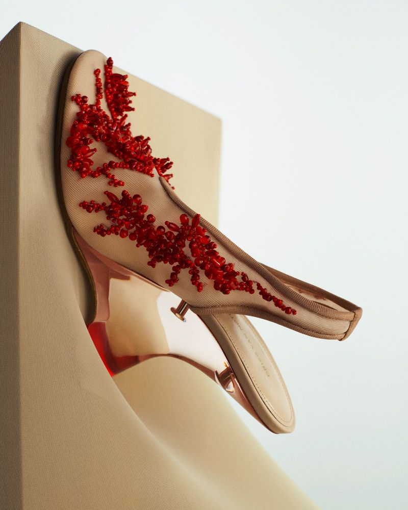 Beige heeled mule with red bead embellishments sits against a curved beige block and white backdrop. Fashionable luxury shoe.