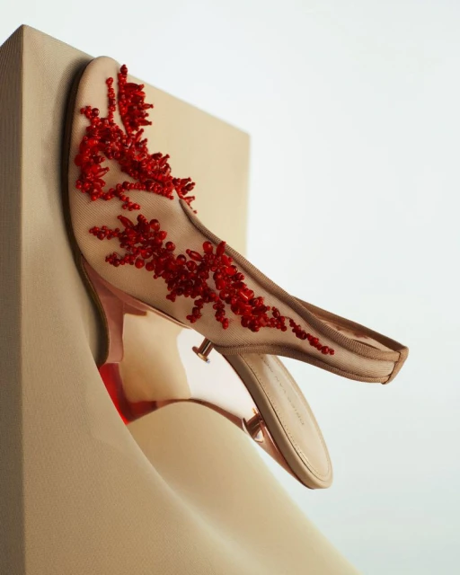 Beige heeled mule with red bead embellishments sits against a curved beige block and white backdrop. Fashionable luxury shoe.
