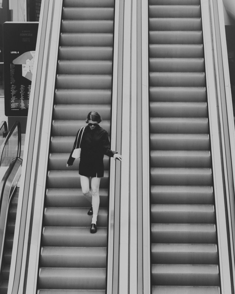 Woman descends an escalator in a minimalist, black and white composition featuring geometric lines, creating a sense of modern solitude.