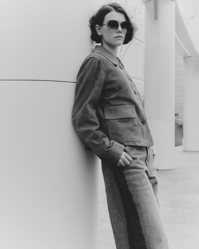 Black and white portrait of a woman in sunglasses, a cropped jacket, and jeans, leaning against a white cylindrical pillar in an urban environment.