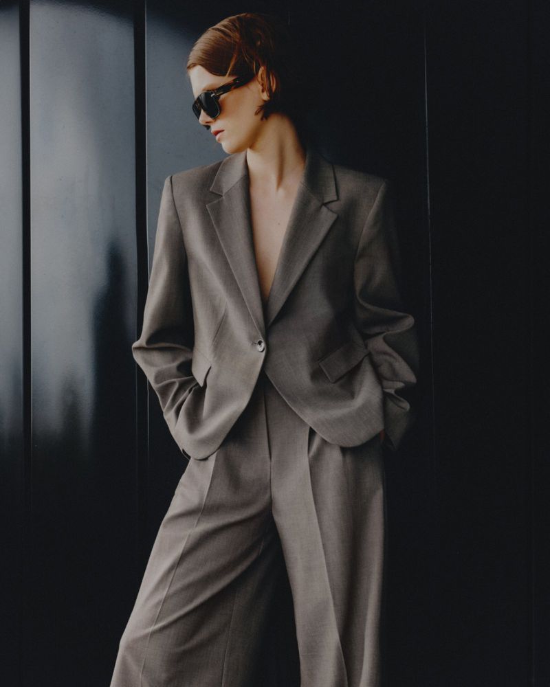 A woman in a stylish taupe suit and sunglasses leans against a dark wall, looking to the side in a fashion editorial shot.