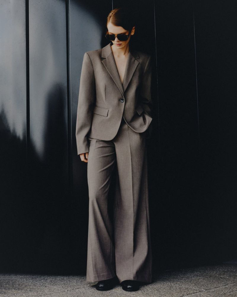 Woman in a grey pantsuit stands against a dark grey panel, accessorized with dark sunglasses, showcasing modern, understated business style.