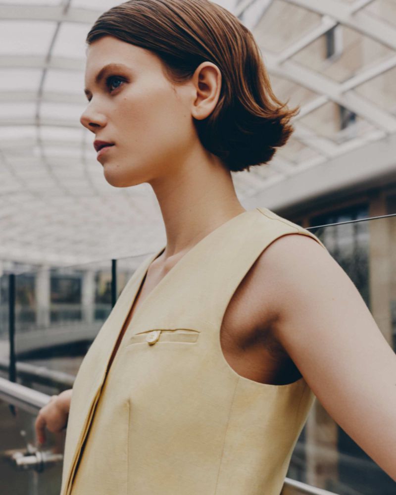 Adult female fashion model in a tan sleeveless jacket poses indoors near a glass railing. Natural lighting, medium shot, neutral expression.
