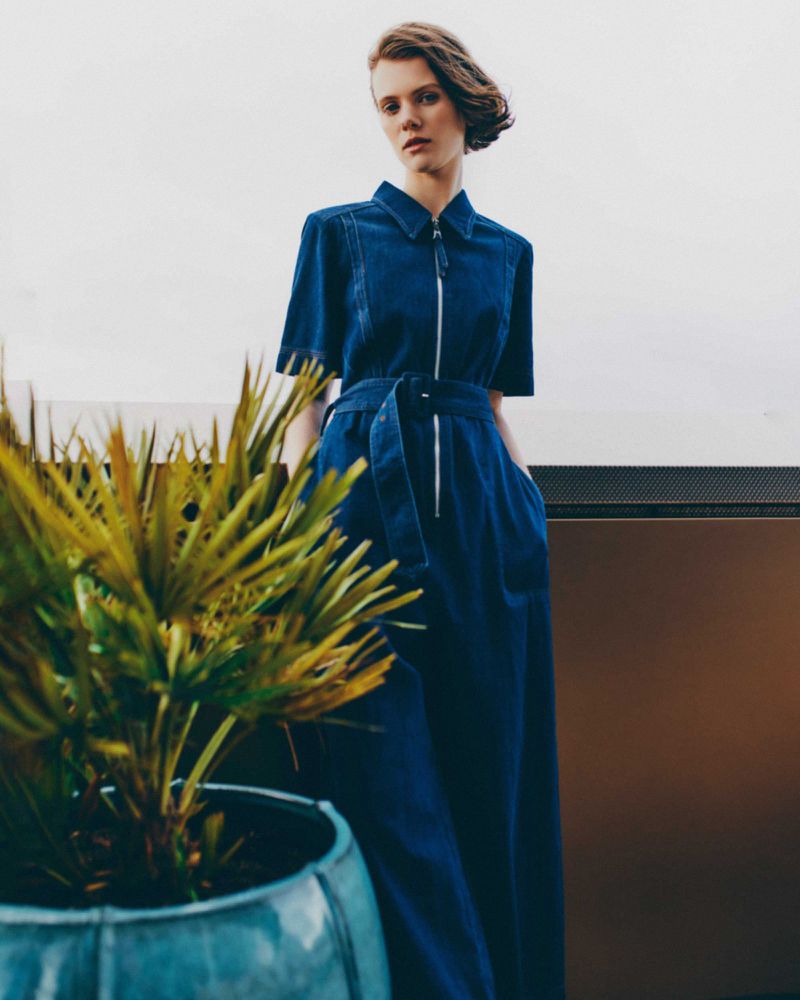 Woman poses outdoors in a blue, zip-front denim jumpsuit with a tie waist. A potted palm is in the foreground.