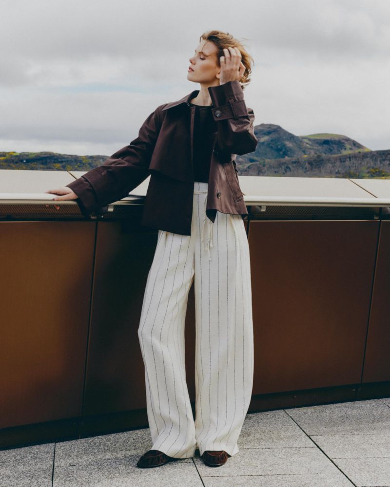 Adult woman poses on a rooftop in a brown jacket and pinstriped wide-leg pants, with a blurred background of mountains and cloudy skies.