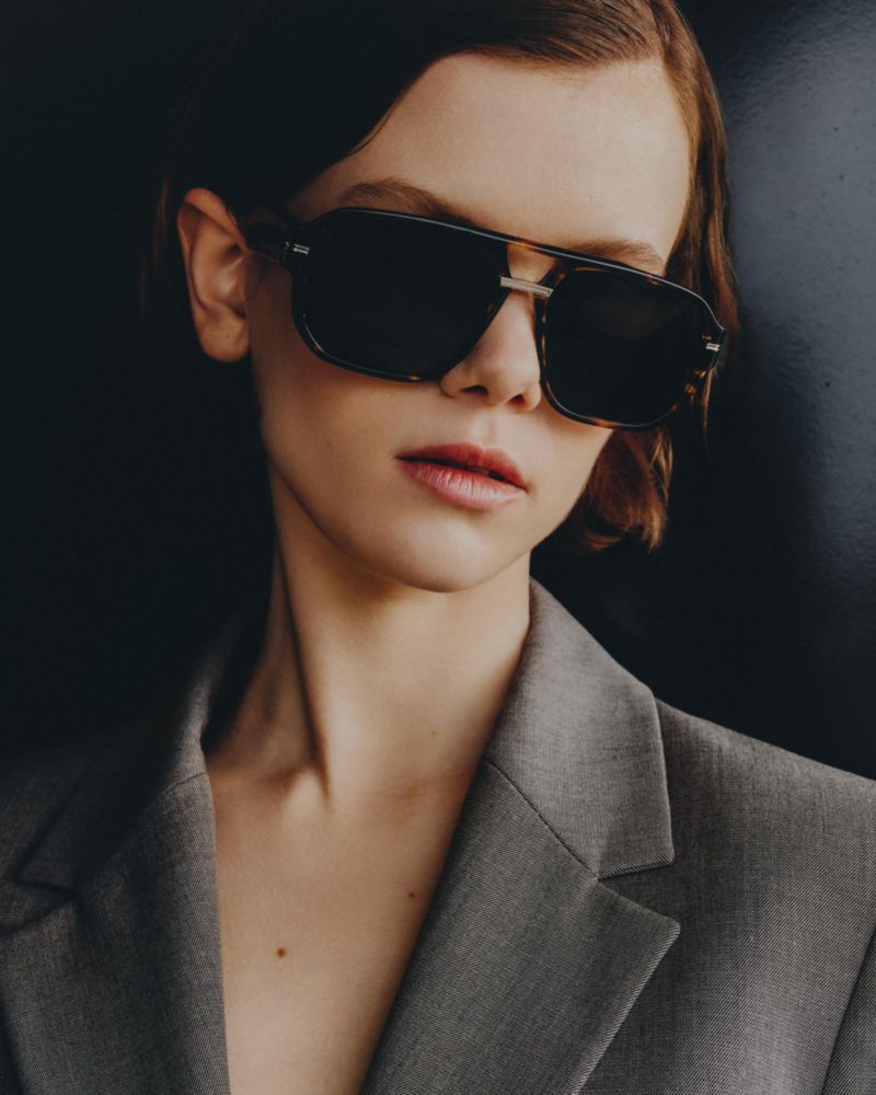 Close-up portrait of a woman wearing aviator sunglasses and a grey blazer against a dark background in a fashion-style photograph.