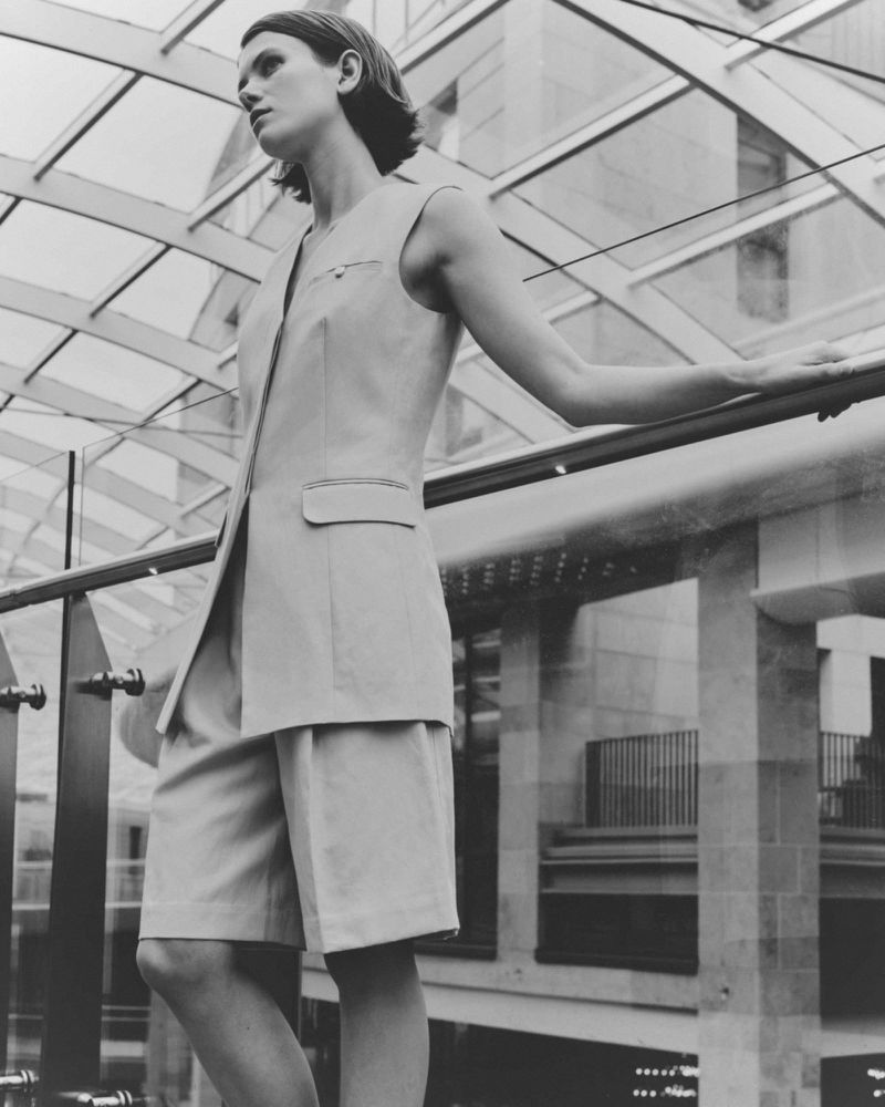 Woman in sleeveless beige suit poses inside building with glass ceiling. Fashion editorial shot with natural light in monochrome.