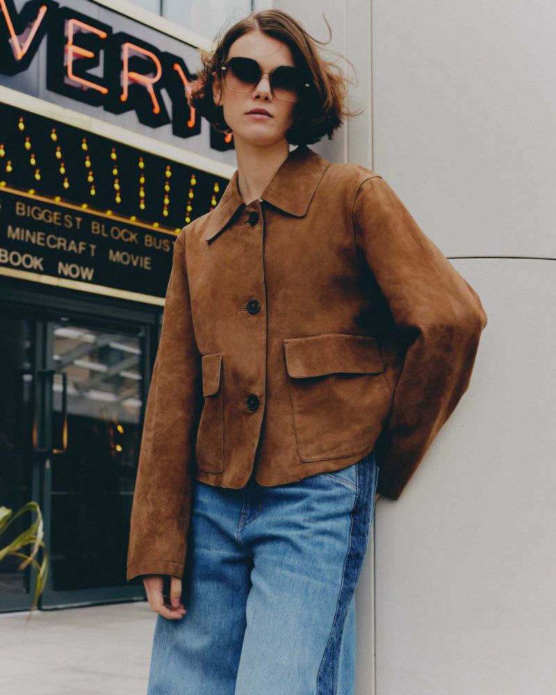 An adult female poses in a brown suede jacket and wide-leg jeans outside of a movie theatre with bright signage, styled with sunglasses.