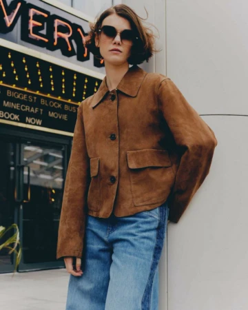 An adult female poses in a brown suede jacket and wide-leg jeans outside of a movie theatre with bright signage, styled with sunglasses.