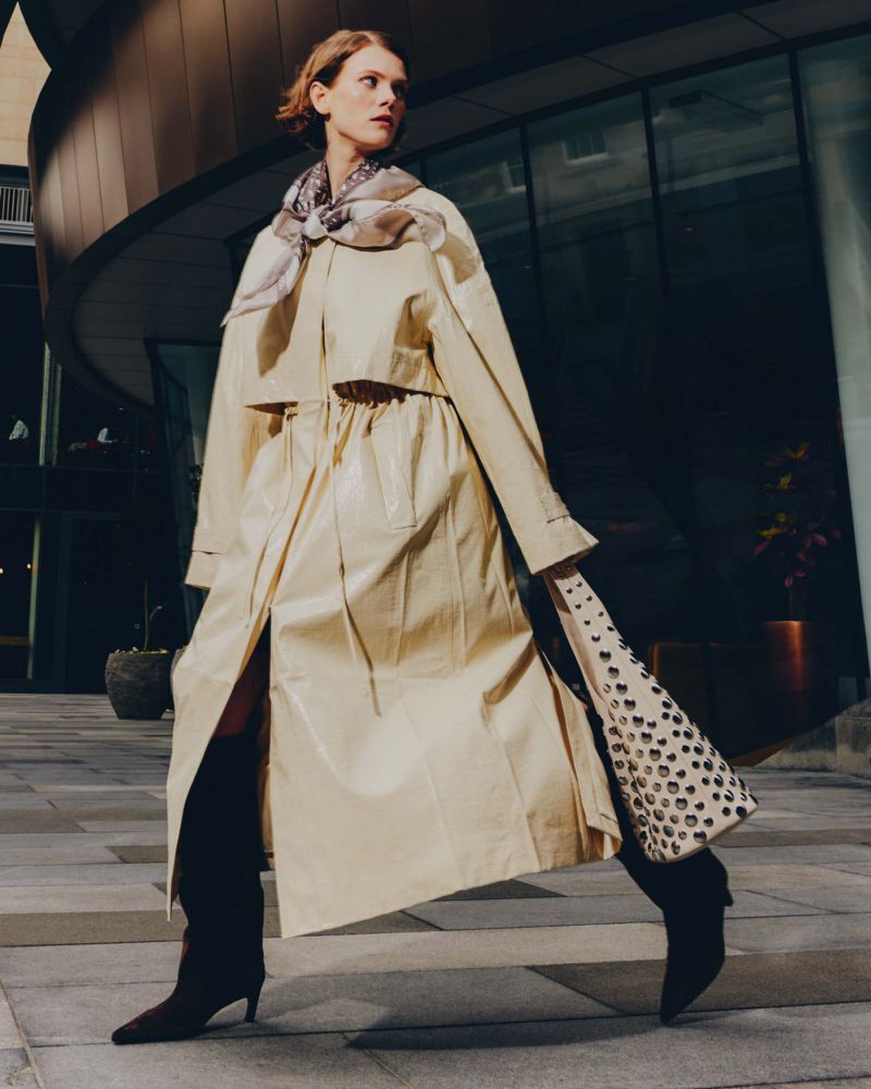A model strides confidently outdoors, showcasing a long beige trench coat, a patterned scarf, and a tote bag with heart-shaped designs.