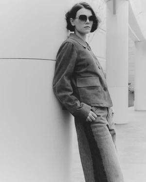 Model in sunglasses leaning against a curved white pillar wearing a buttoned jacket and trousers