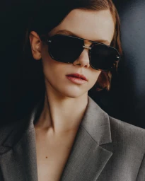 Close-up portrait of a woman wearing dark sunglasses and a grey blazer against a dark background, photographed by Tobias Delcroix.