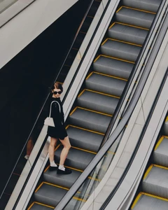 Woman in dark dress and sunglasses standing on a modern indoor escalator holding a white handbag, captured from a high-angle view.