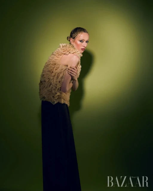 A slender woman in an avant-garde ruffled top and long black skirt poses against a graduated yellow-green background in a studio shot.