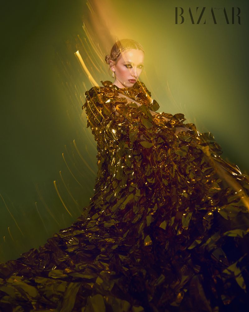 Fashion editorial shot of a woman wearing a shimmering gold dress against a muted green background with visible magazine logo.