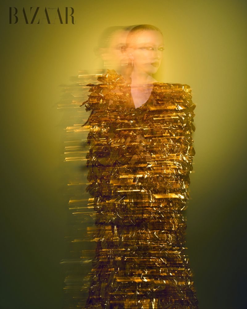 Fashion editorial shot of a woman with blurred motion, wearing a gold dress against a soft, muted gold and green background. Bazaar logo.