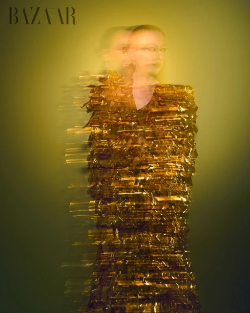 Fashion editorial shot of a woman with blurred motion, wearing a gold dress against a soft, muted gold and green background. Bazaar logo.