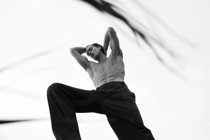 Black and white portrait of a fit adult male in wide-leg pants, posed with arms behind his head against a bright, soft background.