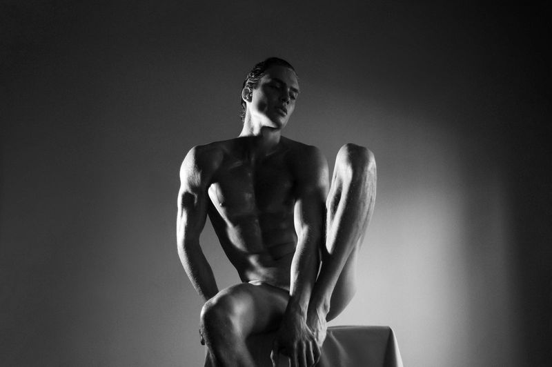 Monochrome portrait of a muscular adult male seated in a studio with dramatic lighting highlighting his physique and anatomy.