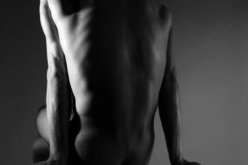 Monochrome nude male figure study: Back view shows muscular physique, veins, and sweat. Dark neutral backdrop with soft, diffused lighting.