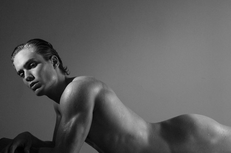 Monochrome portrait of a muscular adult male model with wet hair, lying down indoors in a photo studio with gray background.