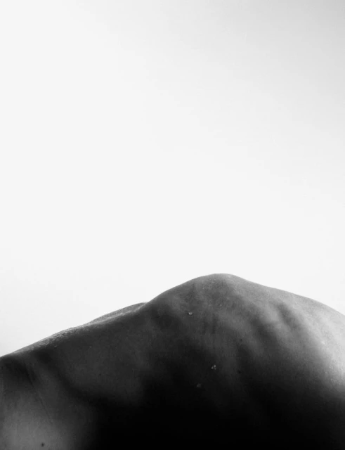 Close-up black and white photograph of the back of a person's shoulder and neck against a plain, bright white background.