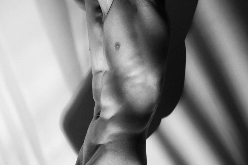 Monochrome studio shot of a muscular adult male torso, showing toned abs and chest, illuminated by bright, patterned lighting.