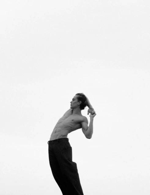 Black-and-white portrait of a muscular adult male with long hair, posing with a bent back against a bright white sky background.