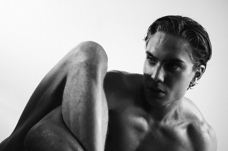 Black and white studio portrait of a shirtless adult male with slicked-back hair, focusing on facial features and body contours in a minimalist style.