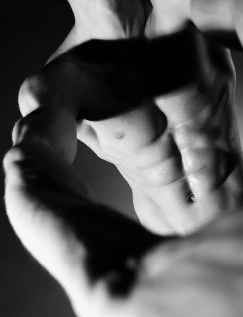 Black and white photo of a muscular man's torso showing abdominal muscles and arms, lit to highlight body contours and muscle tone.