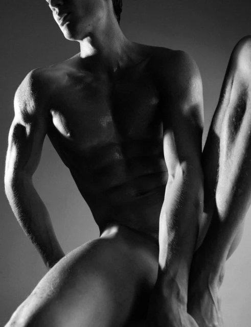 Black and white artistic nude portrait of a male figure with pronounced muscularity in a studio setting against a grey gradient backdrop.