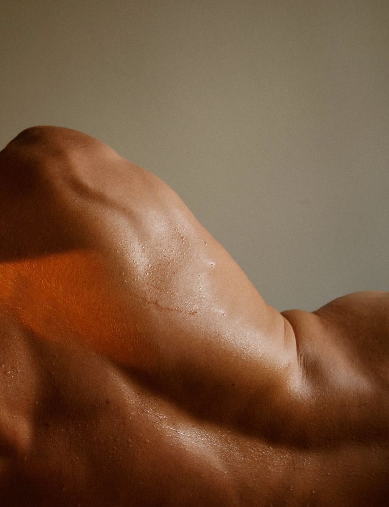 Close-up of a muscular adult male torso and back. The tanned skin is oiled and reflects light, emphasizing muscular definition and tone.