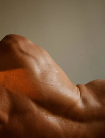 Close-up of a muscular adult male torso and back. The tanned skin is oiled and reflects light, emphasizing muscular definition and tone.