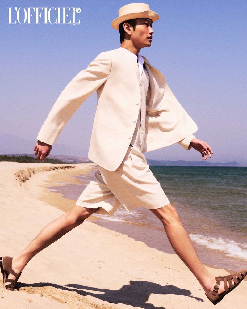 Asian male model in a cream suit, hat, and sandals walks on a beach with the ocean and sky in the background during the daytime.
