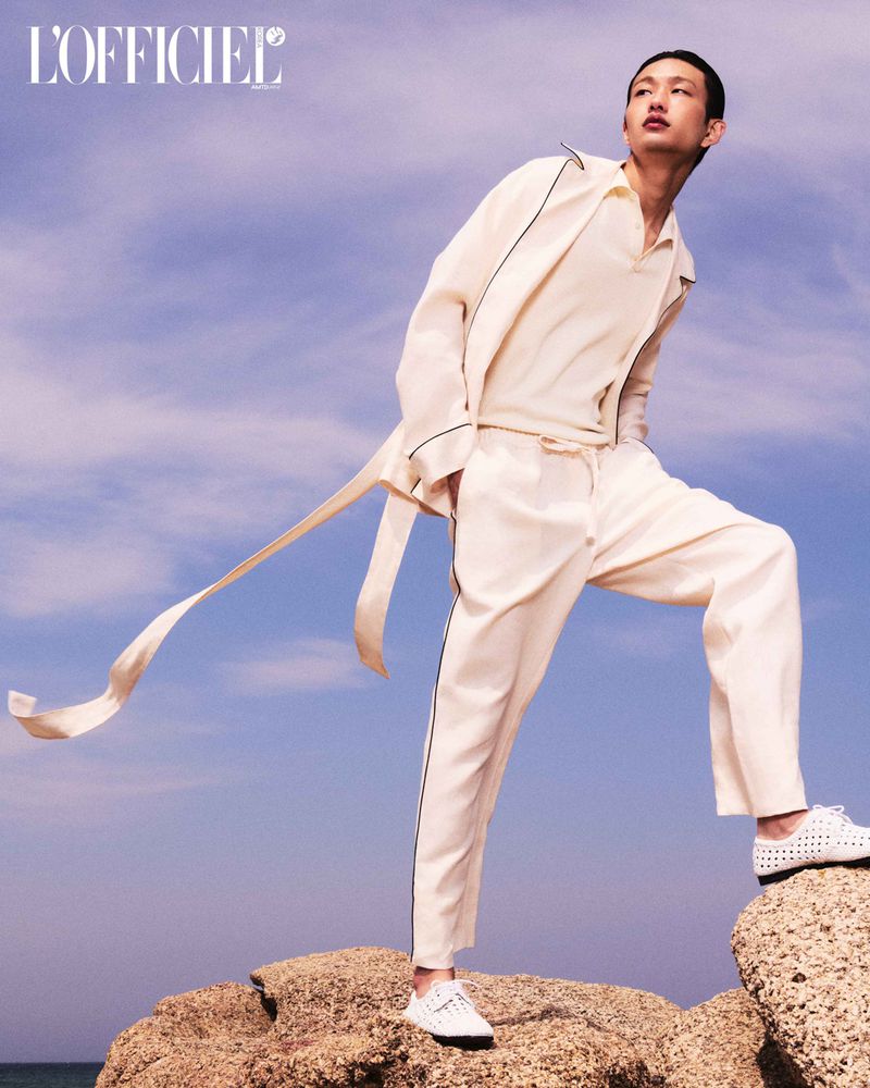 Fashion shot of a male model wearing a cream-colored suit and white shoes posing on rocks against a blue sky. L'Officiel logo visible.